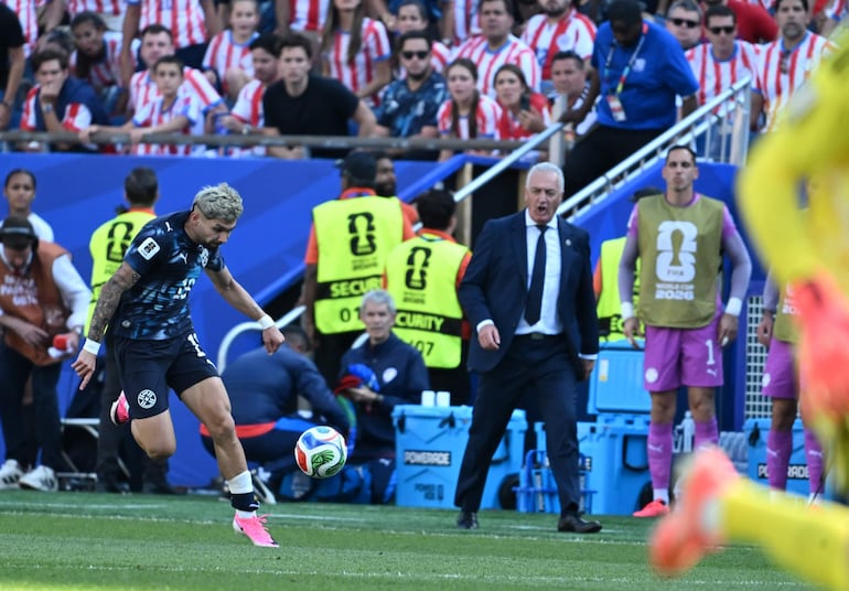 Gustavo Alfaro en traje gesticulando mientras Julio Enciso, con camiseta paraguaya, está a punto de patear el balón en un estadio lleno.