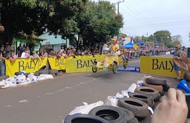 Multitud observa un auto amarillo en la competencia de Autos Locos, mientras sostienen un banner de Baly con entusiasmo y alegría.