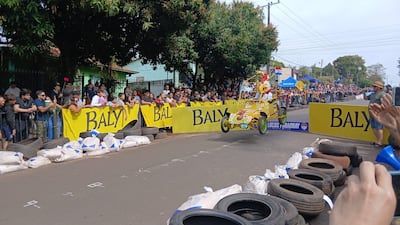 Multitud observa un auto amarillo en la competencia de Autos Locos, mientras sostienen un banner de Baly con entusiasmo y alegría.