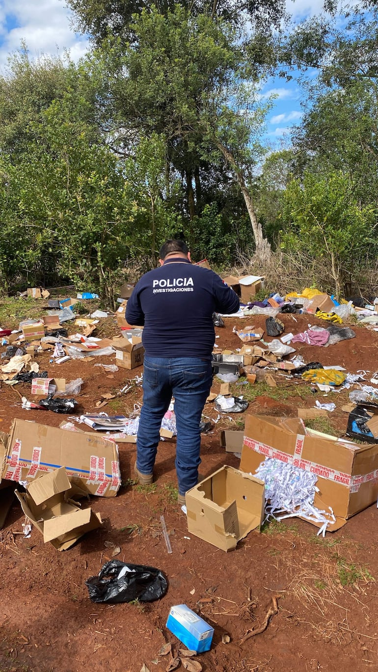 Un hombre con chaqueta de 'POLICÍA INVESTIGACIONES' observa desechos en un terreno al aire libre, rodeado de cajas y plastico.
