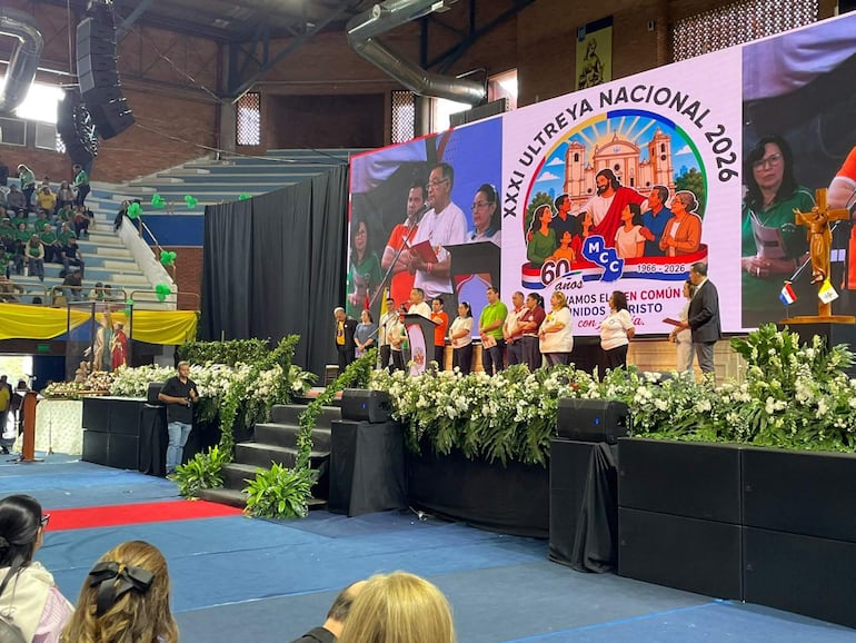 Multitud de personas con camisetas de colores en evento festivo, decorado con flores y un orador en traje en el escenario.