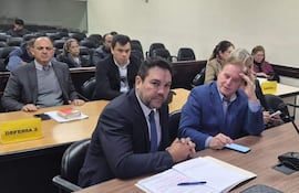 Hombres en trajes, uno con corbata oscura, discutiendo en mesa con documentos en un ambiente cerrado.
