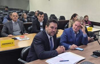 Hombres en trajes, uno con corbata oscura, discutiendo en mesa con documentos en un ambiente cerrado.