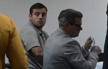 Héctor Iván Grau con camiseta gris observa hacia atrás, mientras un hombre mayor de traje gris sostiene un documento en la mesa.