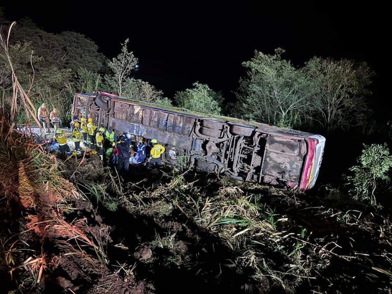 Rescatistas en uniformes amarillos trabajando alrededor de un autobús volcado en un área boscosa, luces de rescate iluminando la escena.