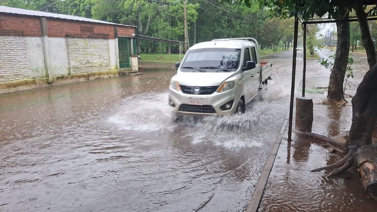 Un vehículo utilitario blanco atraviesa una calle inundada entre paredes de ladrillo y cemento en un entorno residencial