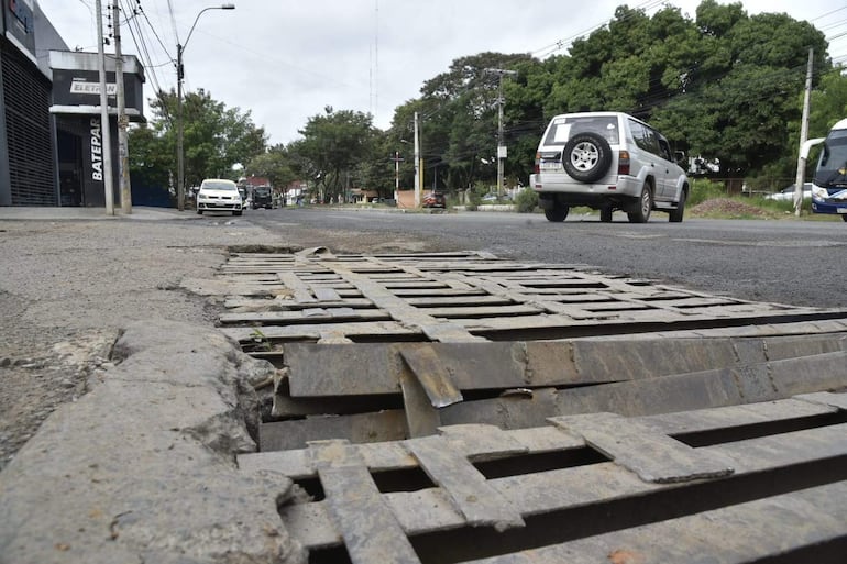 Calle sin personas, con desagüe dañado en primer plano y vehículos estacionados en la vía.