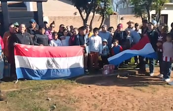 Grupo de personas con banderas de Paraguay en un ambiente rural, escuchando a un hombre con camiseta a rayas que habla en el centro.