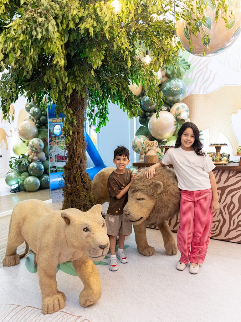 Niño en camiseta marrón y niña en camiseta blanca, sonriendo junto a figuras decorativas de leones y un árbol con globos verdes y dorados.