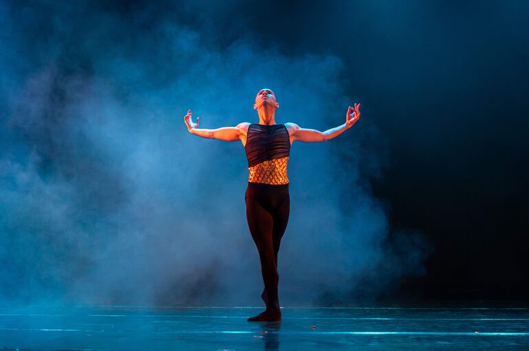 Bailarín masculino con torso descubierto y pantalón negro, en postura expansiva bajo luces tenues y humo en el escenario.