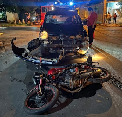 Motocicleta roja volcada y automóvil negro dañado, con espectadores atónitos en un entorno urbano nocturno.