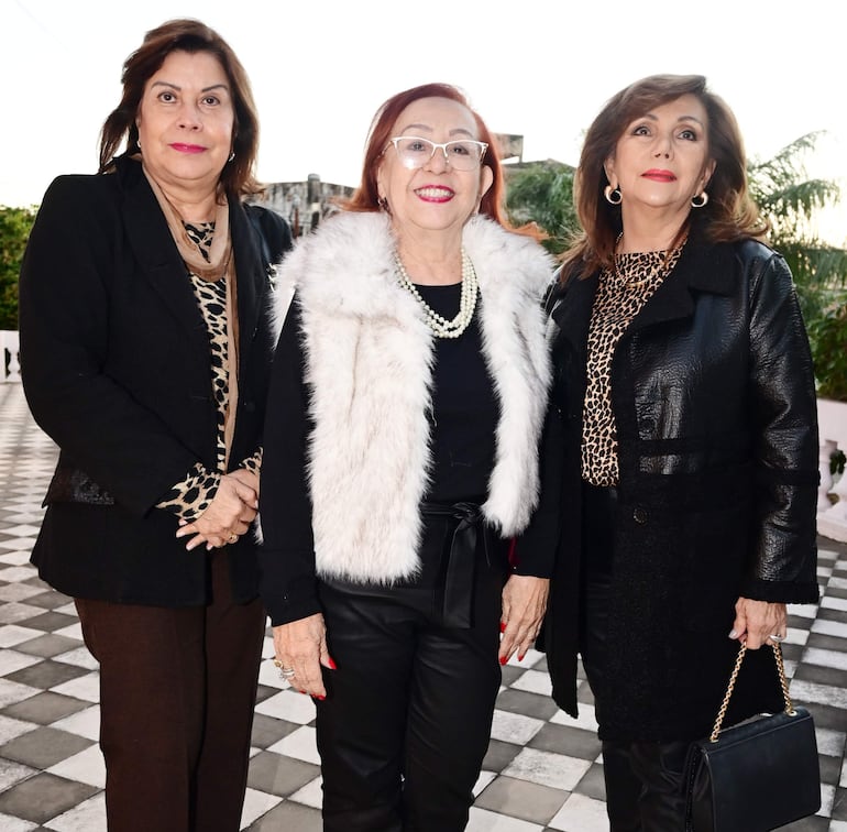 Tres mujeres sonrientes: una con chaleco negro, otra en abrigo blanco y una más con chaqueta negra, sobre piso de baldosas en un entorno exterior.