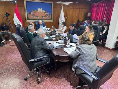 Isaías Ricardo Fretes analizando documentos, mientras José Emilio Argaña Contreras escucha, en un ambiente formal con bandera de Paraguay.