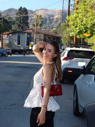 Mujer sonriendo con blusa blanca con lunares, bolso rojo, en la calle con letrero de Hollywood detrás.