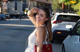 Mujer sonriendo con blusa blanca con lunares, bolso rojo, en la calle con letrero de Hollywood detrás.