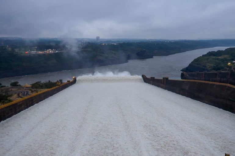 Panorama de la represa de Itaipú con agua fluyendo rápidamente, montañas al fondo y cielo grisáceo.