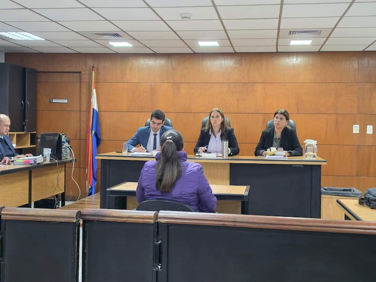 Mujer de espaldas con chaqueta morada testifica ante un escritorio, mientras un hombre y dos mujeres toman notas.