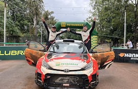 Dos pilotos sonrientes con trajes de carrera celebran en un Toyota sucio en un entorno natural.
