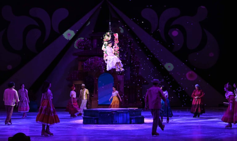 Artistas en trajes vibrantes participan en la producción de Disney on Ice, con luces de colores y una figura femenina en hamaca decorada con flores.