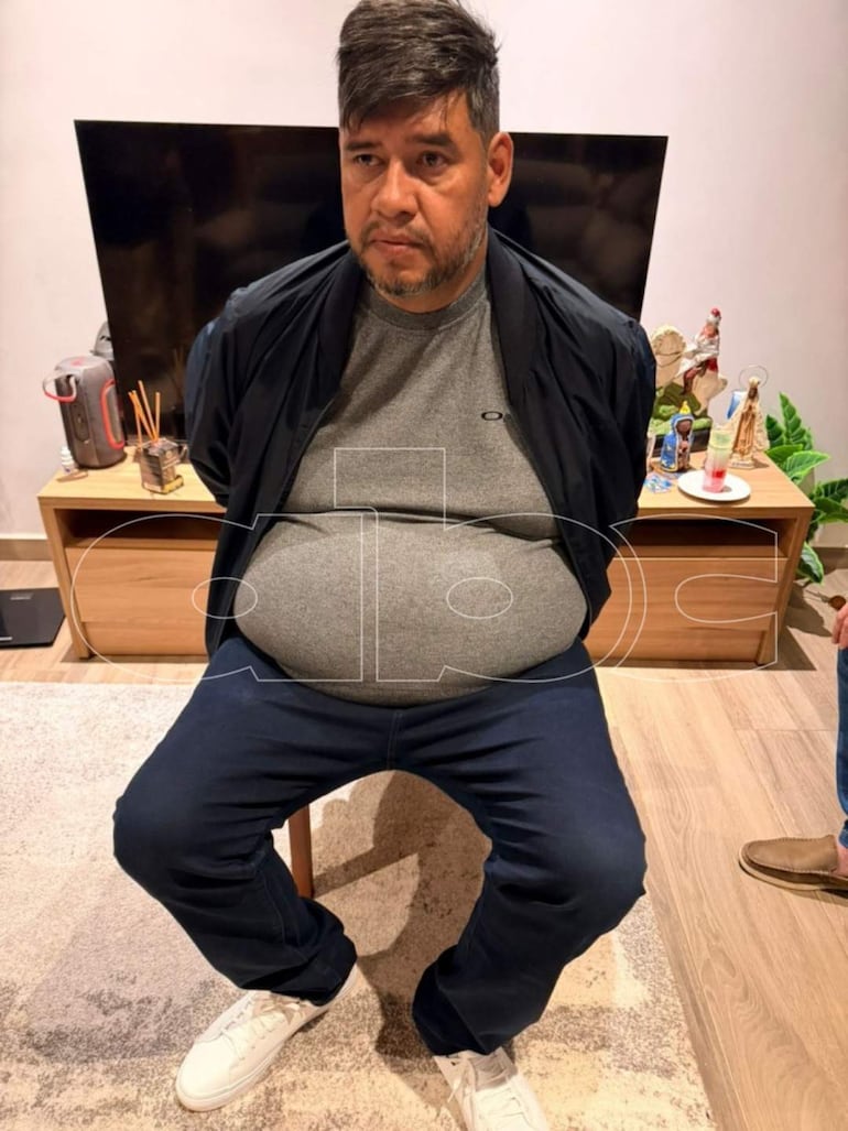 Hombre robusto, Narciso Ayala, sentado en una sala con camiseta gris y abrigo negro, con expresión seria y ambiente privado.