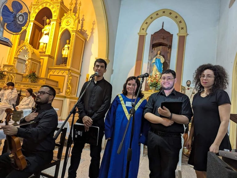 Grupo de cuatro personas en una iglesia. Hombre con violín gesticula mientras dos mujeres sonríen, fondo con altar religioso.