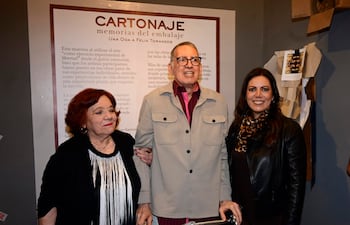 Mujer mayor con cabello rojo sonríe, hombre con andador habla, mujer con abrigo negro escucha en galería de arte.