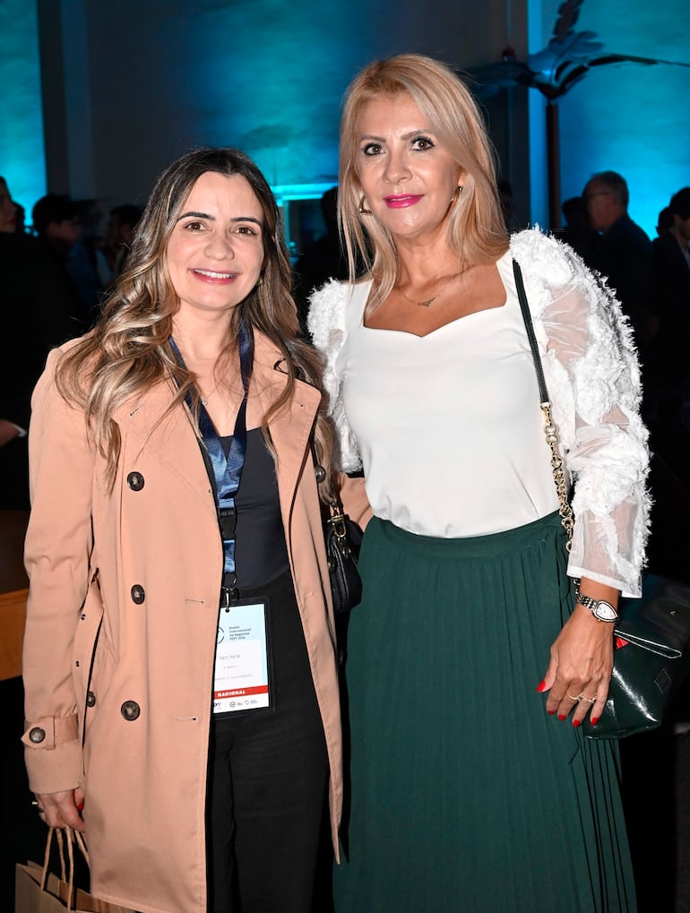 Mujer a la izquierda con abrigo beige e identificación, otra con blusa blanca y falda verde, ambas sonrientes en ambiente formal.