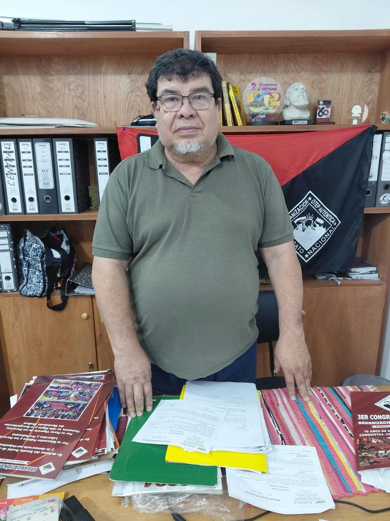 Gabriel Espínola con cabello oscuro y barba canosa, de pie frente a mesa llena de documentos, en ambiente de oficina organizado.