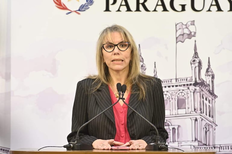 Liz Kramer, con chaqueta a rayas y blusa roja, habla al público desde un micrófono en un auditorio.