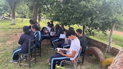 Grupo de estudiantes sentados en sillas, vistiendo pantalones deportivos azules y camisetas, rodeados de naturaleza y árboles.