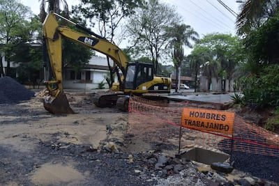 Excavadora amarilla CAT trabajando en una obra pública en un entorno urbano con árboles y edificios alrededor.