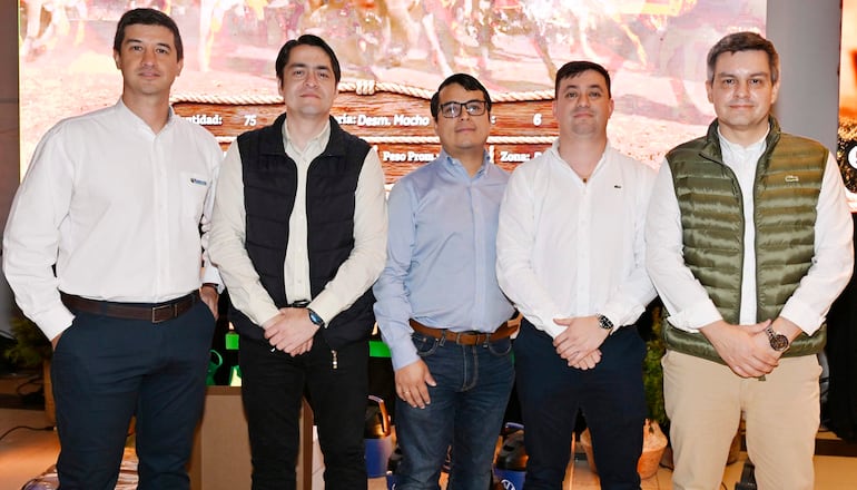 Cinco hombres de pie en un salón de conferencias, vestidos formalmente con camisas y chalecos, mostrando posturas profesionales.
