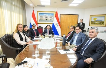 Jorge Ramón Avalos Marinho en traje oscuro, dialoga mientras Hugo Joel Meza sonríe, en reunión con banderas de Paraguay de fondo.