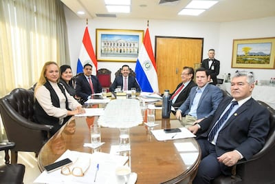 Jorge Ramón Avalos Marinho en traje oscuro, dialoga mientras Hugo Joel Meza sonríe, en reunión con banderas de Paraguay de fondo.