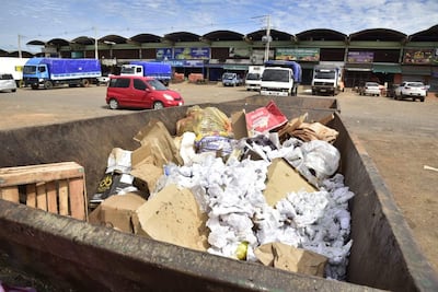 Contenedor de basura lleno de desechos, con vehículos estacionados en un área de carga y descarga en un mercado.
