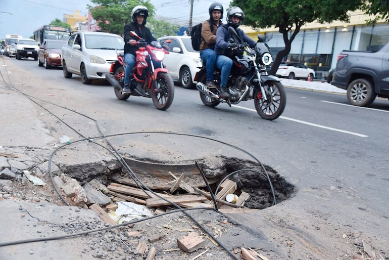 Motociclistas con cascos frente a un bache cubierto de escombros en una calle congestionada, rodeados de vehículos en espera.