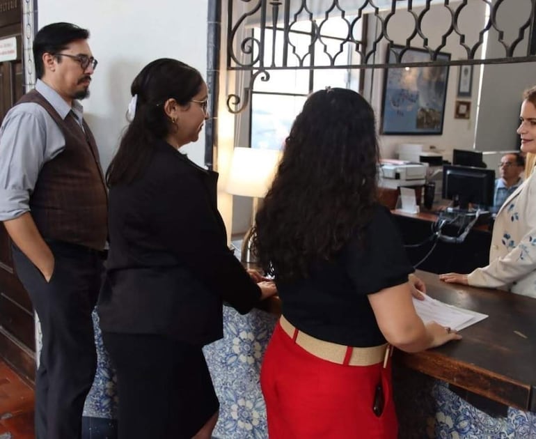 Mujer de cabello oscuro con blusa negra sostiene documentos, mientras otra mujer clara observa desde un mostrador. Hombres en un ambiente de oficina.