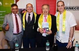 Cuatro hombres sonrientes en trajes formales, dos llevan bufandas amarillas, uno sostiene un libro, en un ambiente interior.