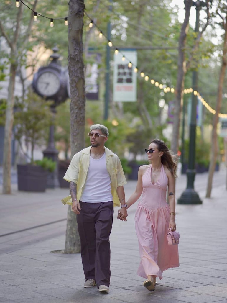 Julio Enciso con camiseta blanca y camisa amarilla, junto a Ramón Sosa en vestido rosa, sonríen caminando de la mano por una calle arbolada.