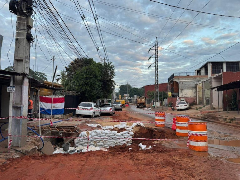 Calle en construcción con sacos de arena y conos de señalización, maquinaria aparcada y bandera de Paraguay en el fondo.