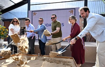 Seis personas, dos en atuendo casual y cuatro en ropa formal, sostienen palas mientras echan arena en ceremonia al aire libre.