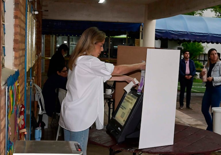 Leticia Ocampos con cabello largo y suelto, vistiendo camisa blanca, interactúa con la máquina de votación en un salón.