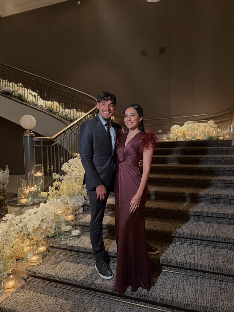 Gustavo Gómez sonríe junto a mujer en vestido marrón con plumas, en elegante escalera decorada con velas y flores.