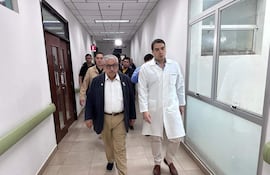 Isaías Fretes, con saco oscuro y camisa blanca, camina junto a Miguel Gómez, en bata blanca, en un pasillo del hospital.