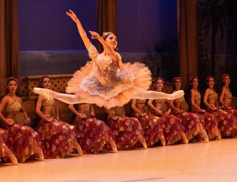 Bailarina en tutú claro realiza salto acrobático, mientras un grupo de bailarinas en trajes tradicionales observa sentadas.