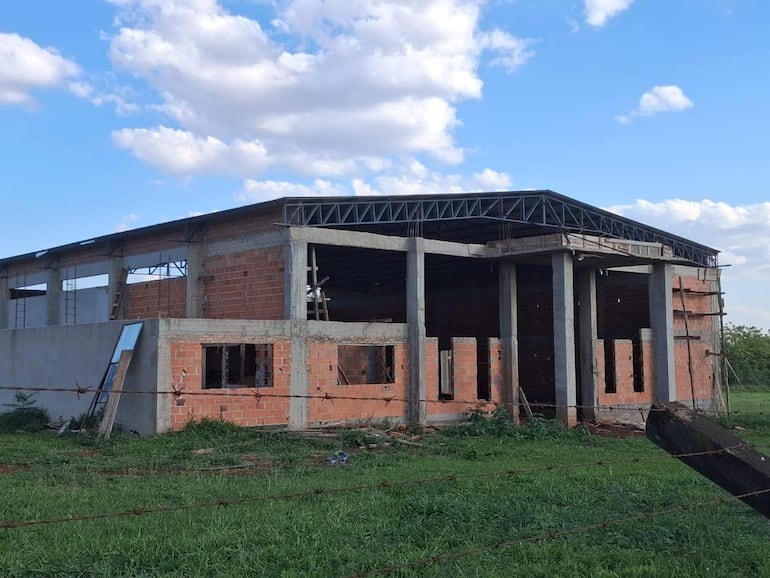 Edificio en construcción con estructura de ladrillo, concreto y techo metálico, rodeado de césped y herramientas de obra.