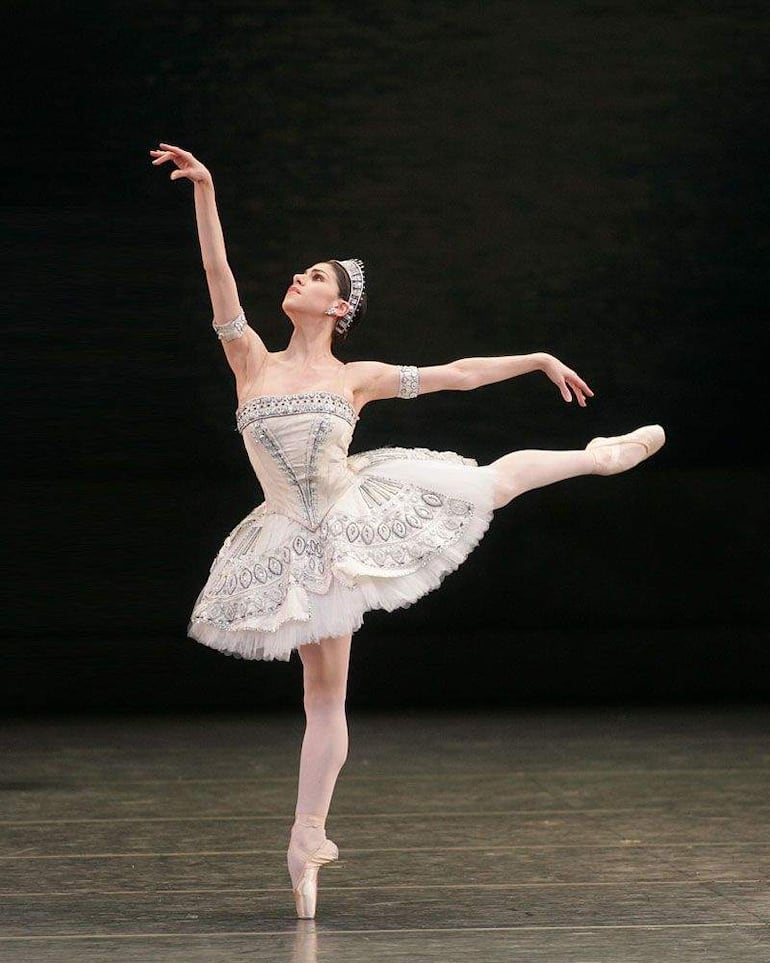 Bailarina en vestido blanco, en primera posición, con brazos extendidos, en un oscuro teatro mientras danza.