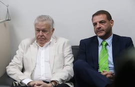 Dos hombres en atuendos formales, uno con chaqueta blanca y el otro con traje oscuro, sentados en un sofá con expresión seria.
