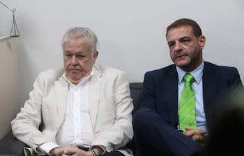 Dos hombres en atuendos formales, uno con chaqueta blanca y el otro con traje oscuro, sentados en un sofá con expresión seria.