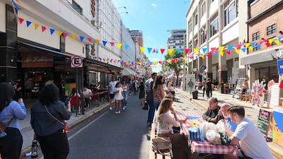 Dos mujeres conversan de pie mientras un grupo de hombres socializa en una calle adornada con banderines coloridos.